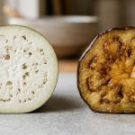 **Alt text:** A side-by-side comparison of a raw, spongy white eggplant slice on the left, and a tender, caramelized cooked eggplant slice on the right, resting on a light-grey stone countertop.