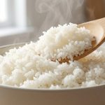 **Alt text:** A close-up of fluffy, steaming white jasmine rice in a matte ceramic bowl, with a wooden paddle lifting a portion to reveal distinct, non-sticky grains in soft, natural light.