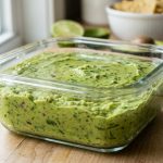 **Alt text:** A close-up of a glass container filled with bright green guacamole, topped with a thin, protective layer of clear water on a sunlit kitchen counter.