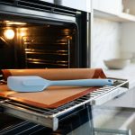 **Alt Text:** A pastel blue silicone spatula and a terracotta-colored baking mat rest on a metal wire rack inside a modern, softly lit oven, with a bright, minimalist kitchen blurred in the background.