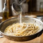 **Alt text:** Close-up shot of glossy spaghetti being tossed in a stainless steel skillet, with a stream of starchy pasta water poured from a ladle to emulsify the creamy sauce. Shot in a bright kitchen with soft, natural light.