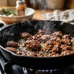 **Alt Text:** A close-up shot of ground beef searing in a hot cast-iron skillet, featuring a caramelized brown crust, sizzling oil, and gentle steam.