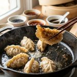 A close-up, high-angle shot of wooden chopsticks lifting a pan-fried dumpling from a seasoned cast-iron skillet, showcasing its crispy, golden-brown lacy crust in a bright, modern kitchen.