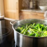 **Alt text:** Vibrant green broccoli, asparagus, and sugar snap peas boiling in an open stainless steel pot on a modern stovetop, with soft steam rising against a warm, blurred kitchen background.