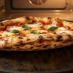 **Alt text:** A close-up shot of a freshly baked pizza on a dark steel baking plate inside a warm oven, highlighting its crispy, charred undercarriage lifting off the steel, topped with melted mozzarella and fresh basil.