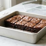 **Alt text:** Fudgy chocolate brownies with a shiny, crinkly crust in a metal baking pan, with an empty glass dish blurred in the background on a marble countertop.