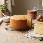 **Alt text:** A freshly baked, golden-brown vanilla sponge cake on a wire cooling rack, with a single slice on a plate showing a fluffy interior, set on a sunlit wooden kitchen counter.
