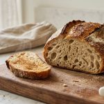 A sliced artisan sourdough bread loaf on a wooden cutting board, showcasing a soft, airy interior with large air pockets. A warm, toasted slice rests nearby on a sunlit kitchen countertop.