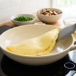 **Alt text:** A perfectly folded, pale yellow French omelet cooking in a cream-colored ceramic skillet. A grey spatula gently lifts the edge of the glossy omelet, set against a bright, sunlit kitchen background.