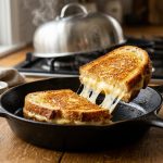 **Alt text:** A golden-brown grilled sourdough cheese sandwich in a cast-iron skillet, sliced in half with a dramatic stretch of melted cheese under warm, natural kitchen light.