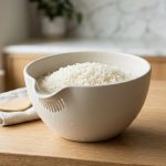 **Alt text:** Raw, glistening white rice in a minimalist off-white Japanese washing bowl with drainage slots, illuminated by soft morning sunlight on a clean kitchen counter.
