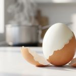 **Alt text:** A close-up of a hard-boiled egg being peeled on a bright kitchen counter, its shell sliding off in a perfect spiral to reveal a flawless, glossy white surface.