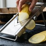 **Alt text:** Close-up of a sleek stainless steel mandoline slicer cutting a yellow potato, with a paper-thin slice gliding off the blade next to a neat fan of uniform potato slices on a dark slate countertop.