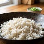 **Alt text:** Macro photograph of fluffy, perfectly separated grains of cooked jasmine rice in a dark carbon steel wok, illuminated by soft natural light with a blurred background of chopped green onions.