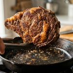 Close-up of a thick ribeye steak searing in a hot cast-iron skillet, showcasing a perfectly caramelized, crispy golden-brown crust with sizzling oil droplets under warm, dramatic lighting.