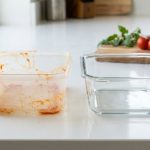 **Alt text:** A side-by-side comparison of two food storage containers on a bright white kitchen countertop: a stained, orange-discolored plastic container on the left, and a sparkling clean, empty glass container on the right.