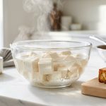 Editorial food photography of fresh white tofu cubes soaking in a steaming glass bowl of water next to crispy, golden-brown pan-seared tofu on a wooden board in a bright, sunlit kitchen.