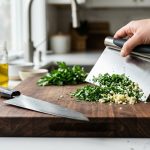 **Alt text:** A close-up of a stainless steel bench scraper scooping chopped herbs and garlic on a wooden cutting board, with a chef’s knife resting nearby in soft morning light.