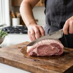 **Alt text:** A close-up of a cook's hands using a chef's knife to score the fat cap of a raw pork chop on a rustic wooden cutting board, with a softly blurred kitchen background.