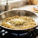 **Alt text:** A close-up, top-down shot of translucent, caramelized diced onions sautéing in a stainless steel skillet, with a fresh mound of minced raw garlic in the center and gentle steam rising.