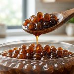 **Alt Text:** A close-up of a wooden spoon scooping glossy, translucent tapioca pearls in rich brown sugar syrup, with a single amber drop falling back into a glass bowl.