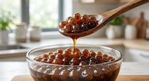 **Alt Text:** A close-up of a wooden spoon scooping glossy, translucent tapioca pearls in rich brown sugar syrup, with a single amber drop falling back into a glass bowl.