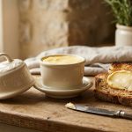 **Alt Text:** An elegant cream-colored ceramic French butter bell sits on a rustic wooden counter next to a slice of toasted sourdough bread with a fresh swipe of butter. The bell’s lid is off, revealing soft golden butter inside, all bathed in warm, soft morning sunlight.