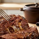 **Alt text:** An extreme close-up of a silver fork pulling apart a tender beef chuck roast, revealing glistening, juicy muscle fibers and melted gelatin under warm light, with a blurred Dutch oven in the background.