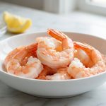 **Alt text:** Plump, glistening cooked shrimp in a white ceramic bowl on a light marble countertop, with a soft-focus background of a lemon wedge and a small bowl of prep ingredients.