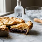 **Alt text:** Close-up of a golden-brown pie crust on a marble counter, with a cut slice showing flaky, airy layers. A cold bottle of vodka and a floured rolling pin are softly blurred in the background.