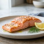 A perfectly cooked, flaky pink salmon fillet glistening on a minimalist ceramic plate, garnished with a lemon wedge and a sprig of dill under soft, natural light.