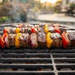 **Alt text:** A close-up of flat stainless steel skewers on an outdoor grill, loaded with juicy, charred beef cubes, red and yellow bell peppers, and red onions, glistening in warm golden hour light.