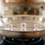 **Alt text:** A clean, macro photograph of perfect spherical water droplets hovering and gliding across the hot surface of a polished stainless steel skillet, demonstrating the Leidenfrost effect.
