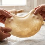 **Alt text:** Close-up of two hands gently stretching warm, elastic dumpling dough on a flour-dusted marble countertop. Faint steam rises in the warm morning light, with folded dumplings in the soft-focus background.