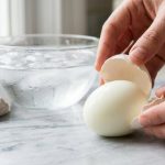**Alt text:** A close-up photograph of a perfect hard-boiled egg being effortlessly peeled on a marble countertop, revealing a flawless, smooth white surface. In the softly blurred background is a glass bowl filled with ice water.
