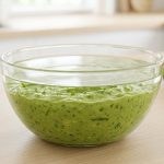 A glass bowl of fresh green guacamole on a light-wood countertop, with a thin, transparent layer of water resting on top that reflects soft kitchen light.