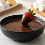 **Alt text:** A fresh strawberry being dipped into a bowl of smooth, glossy melted dark chocolate, with platters of fruit and marshmallows softly blurred in the background.