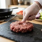 **Alt text:** A close-up of a chef’s hand pressing a shallow dimple into the center of a raw, marbled beef burger patty on a dark slate board, with fresh ingredients blurred in the sunlit background.