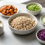 A bright, minimalist food photograph of a healthy grain bowl spread on a light gray tabletop. A central ceramic bowl of fluffy quinoa is surrounded by small dishes of roasted sweet potatoes, green edamame, sliced avocado, and shredded purple cabbage under soft, natural light.