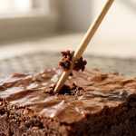 **Alt text:** An extreme close-up of a wooden toothpick being lifted out of a fudgy chocolate brownie, with a few moist crumbs clinging to its center. The brownie features a shiny, crinkled crust and a dense interior.