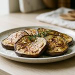 An editorial food photograph of caramelized eggplant slices on a minimalist white plate, garnished with flaky sea salt and a sprig of thyme, in soft, natural light.