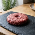 **Alt Text:** A close-up photograph of a raw ground beef burger patty with a shallow thumbprint dimple in the center, resting on a dark slate cutting board in a softly lit kitchen.