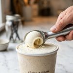 **Alt text:** Close-up of a hand holding a sleek aluminum scoop, effortlessly carving a perfect, smooth curl of vanilla bean ice cream. A subtle glisten of melted cream shows at the scoop's leading edge on a clean white marble countertop.
