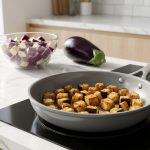 **Alt text:** Golden-brown caramelized eggplant cubes cooking in a modern ceramic skillet on an induction stovetop, with raw purple eggplant in the bright, sunlit kitchen background.