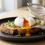 **Alt Text:** A perfectly poached egg, sliced open to reveal a runny golden yolk, resting on a slice of toasted sourdough bread on a ceramic plate. Garnished with cracked black pepper and microgreens in soft morning light.