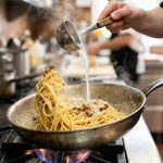 **Alt text:** A close-up photograph of steaming spaghetti being tossed in a stainless steel skillet as a thin stream of pasta water is poured in, creating a glossy, emulsified sauce. Warm, natural lighting highlights the glistening noodles against a softly blurred kitchen background.