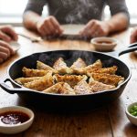 **Alt text:** A cast-iron skillet of steaming gyoza dumplings with crispy, lacy bottoms on a wooden table, surrounded by dipping sauces and the blurred hands of friends.