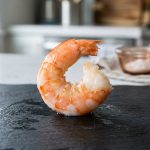 **Alt text:** Macro photograph of a single, plump cooked jumbo shrimp curled on a dark slate plate, glistening under soft natural light with a softly blurred kitchen background.