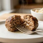 An extreme close-up of a steaming gourmet meatball split open with a silver fork on a minimalist plate, revealing a moist, tender interior. In the soft-focus background, a small bowl holds bread soaking in milk.