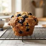 **Alt text:** A close-up photograph of a freshly baked blueberry muffin with a tall, golden-brown sugary crust, resting on a wire cooling rack on a white marble countertop in a bright, sunlit kitchen.
